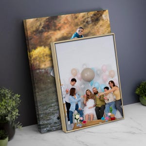 May include: Two framed canvas prints. One shows a family gathering with balloons, the other a landscape with a child. The frames have a gold finish. The family photo includes people toasting with drinks.