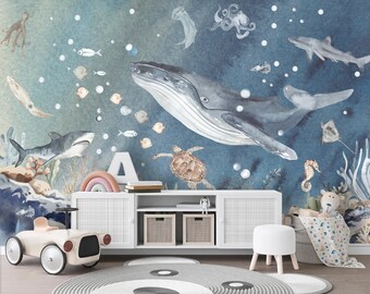 May include: A whimsical nursery scene featuring a blue watercolor mural with marine life, including a whale, sharks, and a sea turtle. A white cabinet holds toys and books, with a toy car in the foreground. A rainbow arch and a white stool complete the decor.