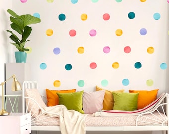 May include: A white metal daybed with colorful throw pillows in orange, green, yellow, and pink. The wall behind the bed is decorated with large, watercolor-style polka dots in various colors. A potted plant and a gold desk lamp are also visible.