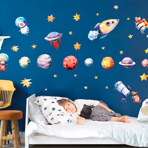 May include: A blue wall with space-themed wall decals featuring planets, stars, a rocket ship, and cartoon dogs wearing astronaut helmets. A child is lying on a white bed with a white and black star patterned pillow.