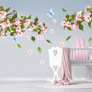 Op de afbeelding: Een wit wiegje met een roze konijnenkussen en een roze deken. De muur achter de wieg is versierd met een bloemenontwerp met roze bloemen, groene bladeren en blauwe vlinders.