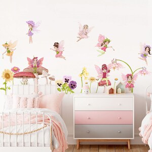 May include: A whimsical bedroom scene featuring fairy wall decals. The fairies, in various poses, are adorned with colorful wings and dresses. The room includes a white dresser with pink and gray drawers, a white bed frame, and floral accents, creating a magical atmosphere.