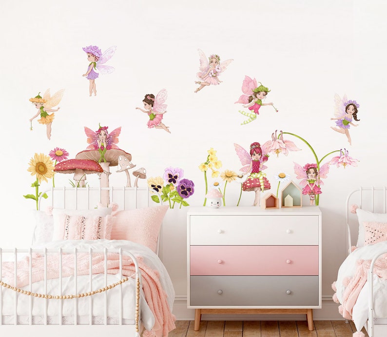 May include: A whimsical bedroom scene featuring fairy wall decals. The fairies, in various poses, are adorned with colorful wings and dresses. The room includes a white dresser with pink and gray drawers, a white bed frame, and floral accents, creating a magical atmosphere.