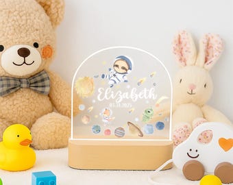 May include: A personalized night light with a space theme, featuring a sloth astronaut, planets, and the name "Elizabeth" with a date. The light sits on a wooden base, surrounded by plush toys and colorful blocks.