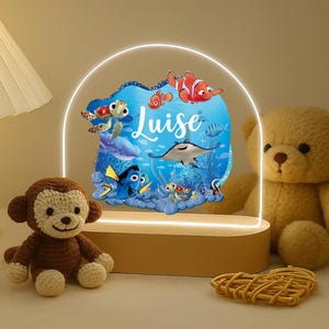 Peut inclure: Une veilleuse décorative avec une scène océanique bleue, des poissons de dessin animé et le nom "Luise". La lumière a une base en bois et une arche lumineuse. Un singe en peluche et un ours en peluche sont au premier plan.