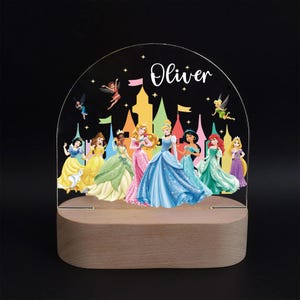 May include: A decorative night light featuring a wooden base and a clear acrylic top. The acrylic displays a colorful illustration of Disney princesses in front of a castle, with the name "Oliver" written above. Fairies and stars are also included.