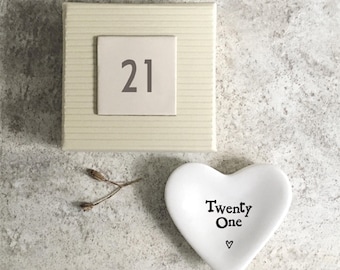 May include: A cream-colored gift box with the number "21" on a square tile, and a white heart-shaped dish with the words "Twenty One" and a small heart symbol. A sprig of dried plant material is also present.