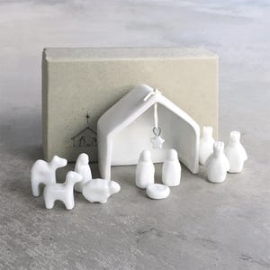 Pode incluir: Um presépio de cerâmica branca com um estábulo, uma estrela e figuras de animais e pessoas. O conjunto inclui uma caixa com um desenho de um estábulo. As figuras são de design simples e moderno.