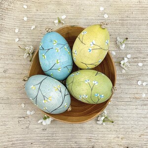 May include: Four painted Easter eggs in a wooden bowl. The eggs are light blue, yellow, and green, each decorated with a branch design and small white and yellow flowers. The bowl sits on a wooden surface scattered with white flower petals.