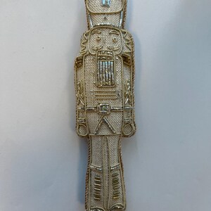 May include: A decorative Christmas ornament in the shape of a nutcracker soldier. The ornament is beige with gold-colored accents, including trim and embellishments. It has a loop at the top for hanging.