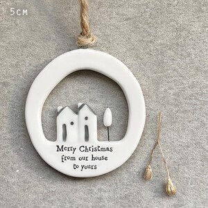 May include: White ceramic Christmas ornament with a circular frame, featuring two small houses and a tree. The text "Merry Christmas from our house to yours" is printed below the houses. The ornament is 5 cm in diameter and hangs from a twine loop.