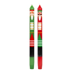 Pode incluir: Duas velas decorativas com desenhos de quebra-nozes. Uma vela é verde com detalhes vermelhos e brancos, a outra é vermelha com detalhes verdes e brancos. Cada vela apresenta um rosto de quebra-nozes e um padrão geométrico.