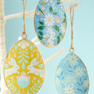 May include: Three decorative Easter egg ornaments with floral and bunny designs. The eggs are in shades of yellow and blue, with gold trim and twine hangers. One egg has the text "Happy Easter".