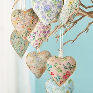 May include: Heart-shaped floral ornaments hanging from a branch. The ornaments display a variety of patterns, including blue hydrangeas, pink roses, and daisies, with gold edging and white ribbon. Each ornament is approximately 10 cm wide.