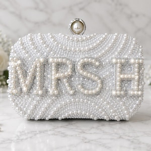 Könnte beinhalten: Eine rechteckige Clutch, die mit weißen Perlen in wirbelnden Mustern bedeckt ist. Auf der Vorderseite befinden sich die Buchstaben MRSH in großen, perlenbesetzten Blockbuchstaben. Der Verschluss ist ein silberfarbener Knopf mit einer Perle.
