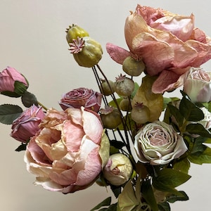 May include: An artificial flower arrangement with large, open roses in pink and peach hues, complemented by green foliage and seed pods. The bouquet includes buds and fully bloomed flowers, creating a naturalistic appearance.