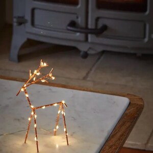 Op de afbeelding: Een koperdraad rendier sculptuur met warm witte LED-verlichting op een marmeren tafel. Een open haard met brandend vuur is op de achtergrond. De tafel heeft een houten rand. Een feestelijke decoratie.