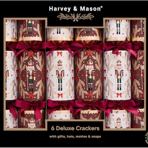 May include: A box of 6 Harvey & Mason deluxe crackers featuring a nutcracker design in white, dark red, and gold. The crackers are tied with dark red ribbons and decorated with gold pine branch patterns, small red stars, and red bows.