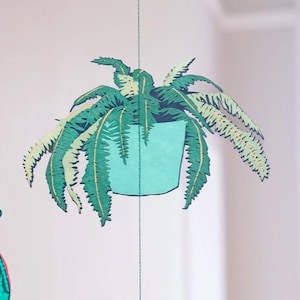 May include: A hanging mobile featuring a paper cutout of a potted fern. The fern has green and light green leaves, and the pot is a light blue. The mobile is suspended by a thin string, creating a whimsical, decorative element.