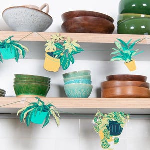 May include: A kitchen shelf displays various bowls in shades of green, brown, and blue. Decorative paper cutouts of potted plants hang from the shelf. The bowls are ceramic and wooden, with varying textures and sizes.