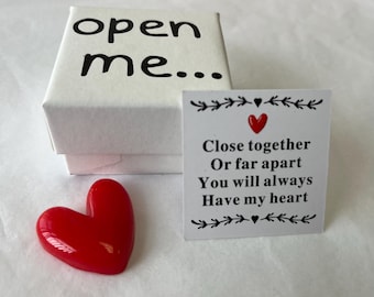 May include: A white gift box with the words "open me..." in black. A red heart-shaped object is next to the box. A small card with a red heart and the text "Close together Or far apart You will always Have my heart."