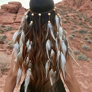 Puede incluir: Una diadema marrón con cuentas de madera y plumas colgantes. Las plumas son blancas y marrones, y están unidas a la diadema con una cuerda marrón. El cabello es largo y ondulado.