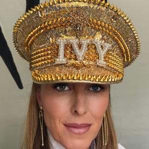 Op de afbeelding: Een gouden hoed met pailletten en het woord "IVY" in witte, parelachtige letters. De hoed heeft een stekelige rand en ingewikkelde gouden versieringen. Een opvallend accessoire voor speciale gelegenheden.