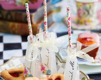 May include: A charming tea party setting with floral-patterned straws in small bottles tied with ribbons. Heart-shaped cookies with jam filling and a "Drink Me" tag are on a decorative tray. A cup and cake are in the background.