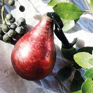 May include: A red glass bottle in a pear shape, with a dark velvet strap. The bottle has a metallic finish and is placed on white paper, alongside a sprig of green leaves and a cluster of dark berries. Likely a decorative item.