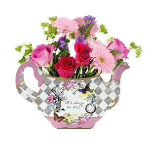 Puede incluir: Una maceta de cerámica con forma de tetera blanca con un borde rosa y un patrón de cuadros blanco y negro. La maceta presenta un diseño floral con una cita que dice "Siempre es hora del té". La maceta está llena de rosas rosas y rojas y margaritas rosas.