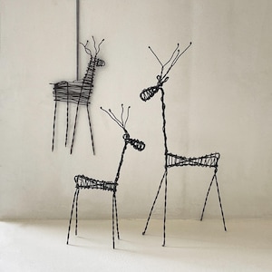 May include: Three black wire reindeer sculptures with detailed antlers and slender legs. The reindeer are different sizes and positioned against a neutral background, creating a minimalist and artistic display. The wire reindeer are a unique home decor item.