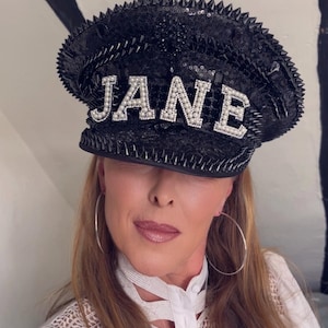 以下が含まれることがあります： 白い文字で「JANE」と書かれた黒いスパンコールの帽子。帽子の縁には銀色のスパイクが付いています。画像内の人物は白いシャツと大きなシルバーのフープイヤリングを着用しています。