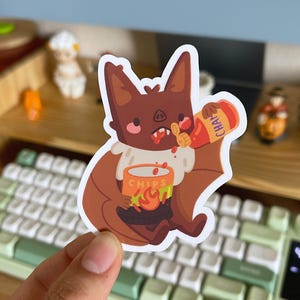 May include: A die-cut sticker featuring a brown bat with pink cheeks and a white ruff around its neck. The bat is holding an orange container labeled CHIPS with a flame graphic and is squeezing a red bottle of hot sauce into its open mouth.