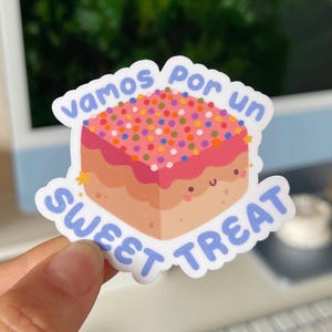May include: A sticker with a cartoon cake with pink frosting and colorful sprinkles. The cake has a happy face and says "vamos por un sweet treat" in blue text.