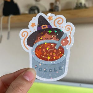 May include: A holographic sticker featuring a character wearing a black witch hat, stirring a grey pot filled with red pozole. The sticker includes orange steam swirls, small stars, and the word POZOLE printed in grey letters on the front of the pot.