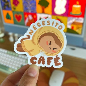 May include: A sticker featuring a cartoon monkey in a yellow hoodie, with the text "NECESITO CAFE" in brown letters. The monkey has a sad expression. The sticker is held in a hand.