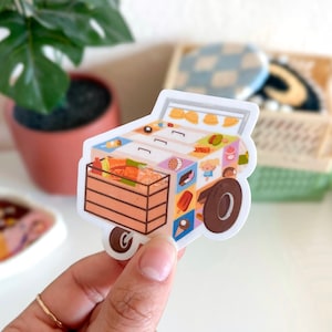 May include: A colorful sticker featuring an illustrated ice cream cart. The cart is filled with various treats, including ice cream cones, popsicles, and pastries. A wooden crate is filled with food. The cart has a brown wheel and a small handle.