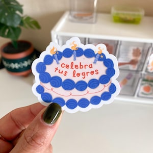 May include: A sticker in the shape of a cake with blue frosting and the Spanish text "celebra tus logros" (celebrate your achievements). The cake has three candles with orange flames. The sticker is held by a hand with a dark green nail.