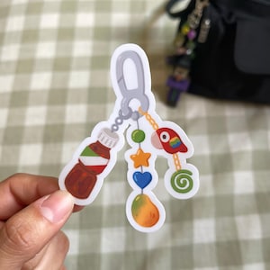 May include: A die-cut sticker featuring a gray carabiner clip attached to three dangling charms: a brown bottle with a white and green label, a vertical string of colorful beads shaped like a star, heart, and fruit, and a red parrot with a green swirl.