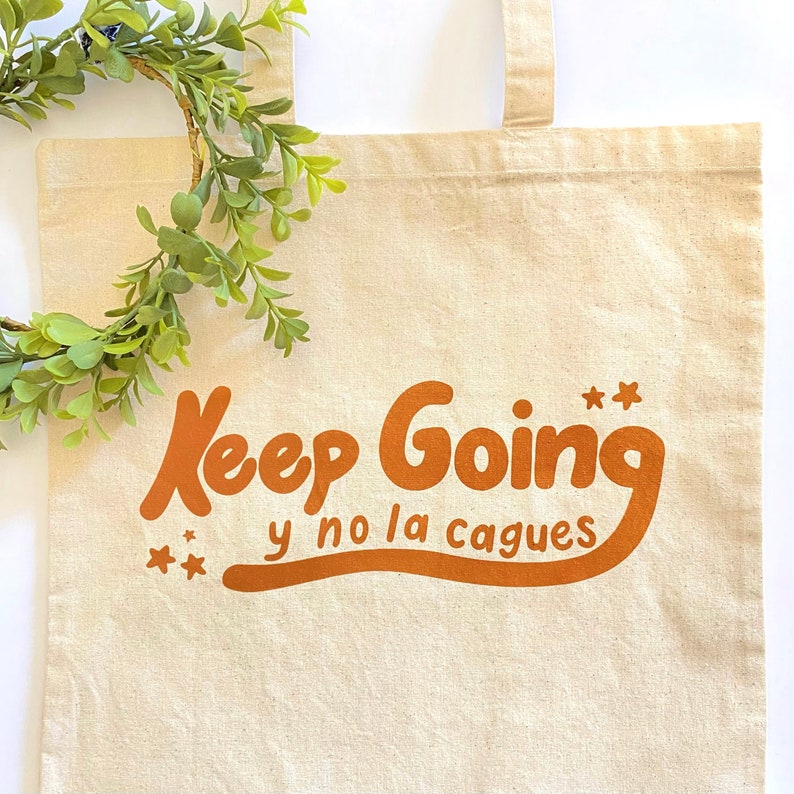May include: A beige canvas tote bag with orange text that reads "Keep Going y no la cagues" with two stars on either side of the text.
