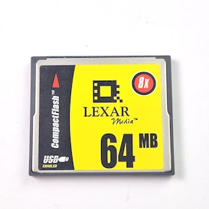 Może przedstawiać: Karta pamięci CompactFlash z żółtą etykietą z logo Lexar Media, napisem "64 MB" i oznaczeniem prędkości "8x". Karta ma czarno-srebrną ramkę i napis "USB Enabled".