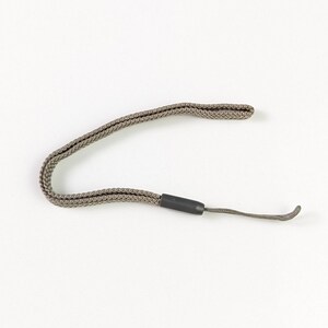 May include: A taupe-coloured woven fabric wrist strap featuring a small, dark grey cylindrical plastic slider. The strap is formed into a loop, with a short, thin cord extension protruding from the slider, designed for attachment to small devices.