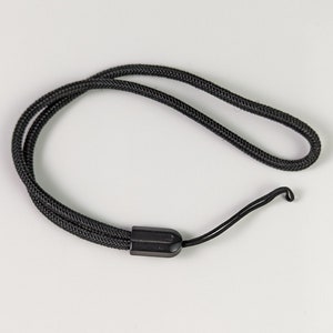 Pode incluir: Uma alça de pulso preta com um laço de cordão trançado e um cordão de fixação fino, unidos por um fecho cilíndrico de plástico preto. O cordão forma um laço contínuo, oferecendo uma maneira segura de transportar pequenos objetos.