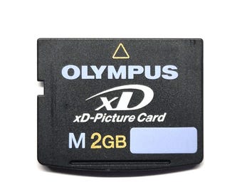 May include: A black Olympus xD-Picture Card with white text reading OLYMPUS, xD, and xD-Picture Card. The memory card features a gold triangle symbol, the letter M, and the text 2GB in gold, along with a small light blue rectangular label area.