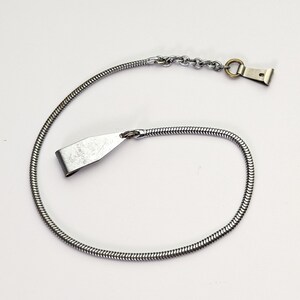Könnte beinhalten: Eine silberfarbene Schlangenkette aus Metall mit einem flachen, spitz zulaufenden Clip an einem Ende und einem kleinen kreisförmigen Ring, der an einem zweiten Clip am anderen Ende befestigt ist. Die Kette hat ein flexibles, abgerundetes Glieddesign.