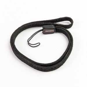 Pode incluir: Uma pulseira de tecido preto com um pequeno conector de plástico retangular com o texto SAMSUNG impresso em branco. A pulseira inclui um laço de cordão preto fino para fixação e uma faixa de tecido plana para prender ao redor do pulso.