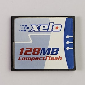 May include: A CompactFlash memory card with a black border. The card features a blue and white design, the brand name "Xelo", and the capacity "128MB CompactFlash" in red text.