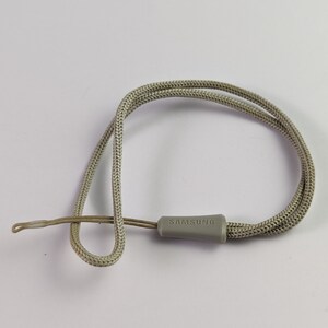 Pode incluir: Uma alça de pulso de cordão trançado cinza-claro com uma pequena peça de conexão de plástico cinza correspondente, gravada com a palavra SAMSUNG. A alça consiste em um laço trançado e um cordão de fixação fino para dispositivos.