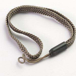 Pode incluir: Um cordão trançado na cor taupe com um conector cilíndrico liso de plástico preto. O cordão é tecido em um padrão plano e flexível que forma um laço, com um segmento de cordão mais fino estendendo-se a partir do componente de plástico preto.