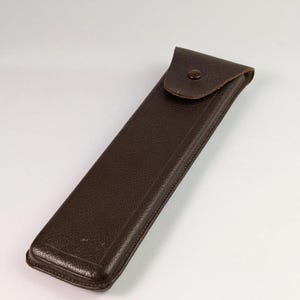 May include: A dark brown rectangular case with a textured surface and a rounded flap closure. The flap is secured by a single dark brown snap button. The case has stitched edges and a slim, elongated profile designed for holding small items.